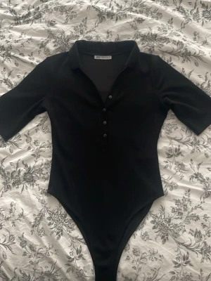 Svart body med krage och knappar - Snygg svart body från NA-KD med klassisk krage och knäppning framtill. Modellen har halvlånga ärmar och en figurnära passform som framhäver siluetten. Perfekt att styla med jeans eller kjol för en trendig look.