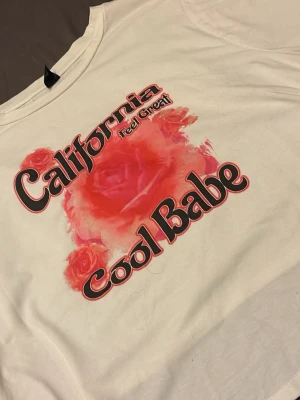 Vit t-shirt California Cool Babe - Vit t-shirt med tryck i rött och svart där det står 'California Cool Babe' och 'Feel Great'. Motiv med röda rosor och färgstänk framtill. Klassisk rund hals och korta ärmar. Perfekt för en avslappnad och trendig look.