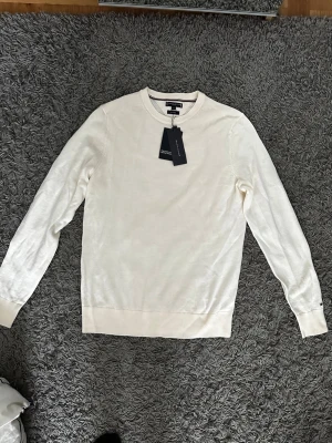 Vit stickad tröja Tommy Hilfiger - Snygg vit stickad tröja från Tommy Hilfiger i mjuk merinoull. Tröjan har rund halsringning, långa ärmar och diskreta ribbade muddar. Perfekt för dig som gillar stilrena och klassiska plagg med en exklusiv känsla.