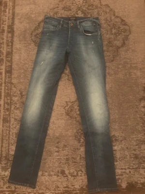 Blå slim fit jeans från Jack & Jones - Säljer ett par blå slim fit jeans från Jack & Jones med snygga slitningar och ljusa tvättade partier på låren och knäna. Klassisk femficksmodell med knappgylf och bälteshällor. Perfekta för en avslappnad och trendig look.