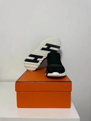  H sneakers med vit sula - Snygga svarta sneakers från H med vit, räfflad sula och stora H-detaljer undertill. Ovandelen är i en mix av mocka och mesh för en sportig och exklusiv look. Klassisk snörning och diskreta dragflikar bak. Perfekta för dig som gillar stilrena och lyxiga sneakers.