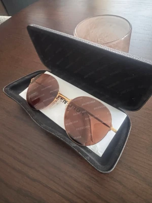 Runda solglasögon från Balenciaga - Säljer ett par riktigt snygga runda solglasögon från Balenciaga med guldiga metallbågar och bruna glas. Glasen har Balenciaga-loggan tryckt över hela ytan för en exklusiv vibe. Kommer med originalfodral och putsduk.