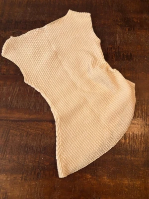 Beige ribbad ärmlös topp - Säljer en vit/beige ribbad topp:) Toppen är ärmlös och har en lätt croppad passform med asymmetrisk nederkant. Perfekt att styla med jeans eller kjol för en trendig look.