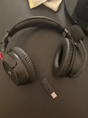 HyperX trådlöst gamingheadset svart - Lite slitet som syns på sista bilden men funkar super. Säljer ett svart trådlöst gamingheadset från HyperX med mjuka over-ear-kuddar och justerbar mikrofon. Headsetet har en stilren design med röda detaljer och medföljer USB-dongel för enkel anslutning till dator eller konsol. Perfekt för gaming och voicechatt.