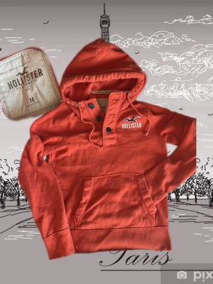 Röd hoodie från Hollister med knappar - Snygg röd hoodie från Hollister med knappar vid halsen och stor magficka. Klassisk logga på bröstet och mjukt, fleecefodrat material. Perfekt för dig som vill ha en casual och bekväm stil. Huva med dragsko för extra komfort.