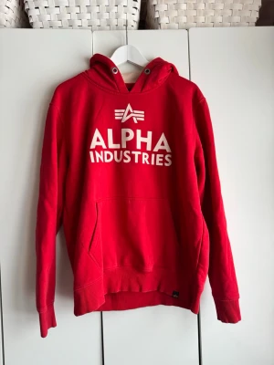 Röd hoodie från Alpha Industries. M.  - Snygg röd hoodie från Alpha Industries med stor vit logga och text på bröstet. Klassisk känguruficka framtill och huva med metallöljetter. Tillverkad i mjukt bomullsmaterial som känns skönt mot huden. Perfekt för en avslappnad streetwear-look. Använd fåtal gånger. Jättefint skick. 
