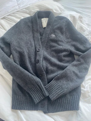 Mörkgrå kofta från Abercrombie & Fitch - Mysig mörkgrå kofta med knappar framtill från Abercrombie & Fitch. Klassisk v-ringad design med ribbade muddar och nederkant. Liten broderad logga på bröstet. Perfekt för lager på lager och chill stil.