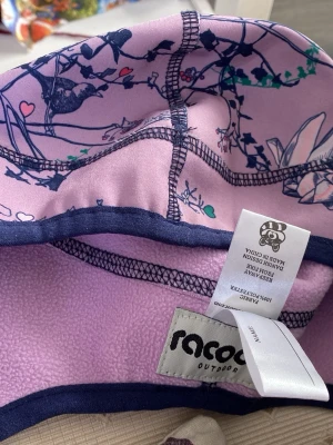 Lila mössa med fågelmönster från Racoon - Säljer en lila mössa från Racoon Outdoor med snyggt fågel- och blommönster i blått, grönt och rosa. Mössan har mörkblå kant och är fodrad med mjuk fleece på insidan för extra värme. Perfekt för dig som vill ha något både stilrent och lekfullt.