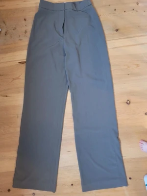 Grå kostymbyxor med rak passform - Snygga grå kostymbyxor med hög midja och rak passform. Byxorna har en klassisk design med dold dragkedja och knapp framtill. Perfekta för en stilren och clean look. Materialet känns mjukt och följsamt, vilket gör dem bekväma att bära hela dagen.