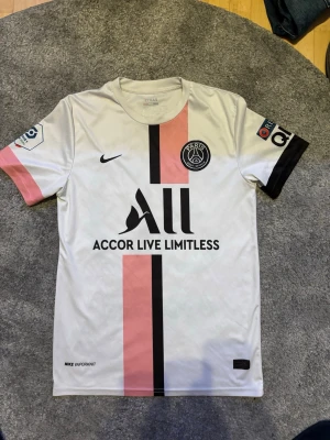 PSG matchtröja vit rosa Nike - Säljer en vit PSG matchtröja från Nike med rosa och svart vertikal rand på framsidan. Tröjan har Paris Saint-Germain-logga på bröstet, sponsortryck och rund hals. Tillverkad i lätt och ventilerande material, perfekt för fotboll eller träning.