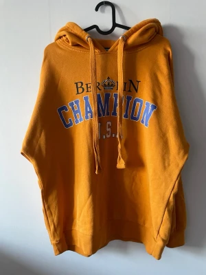 Gul hoodie med tryck Berlin Champion - Snygg gul hoodie från Champion med trycket 'Berlin Champion U.S.A' i blått och svart framtill. Hoodien har huva med dragsko och ribbade muddar. Mjukt material står att det är storlek XS men skulle säga att den är mer en S eller M.