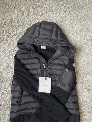 Svart Moncler cardiganjacka med huva - Snygg svart cardiganjacka från Moncler med quiltad front och stickade ärmar. Jackan har huva, dragkedja framtill, fickor med dragkedja och en Moncler-logga på ärmen. Materialmix av dun och stickat ger en modern look.