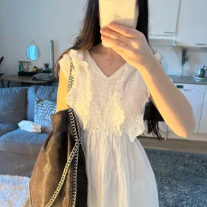 Vit långklänning med volangdetaljer - Säljer en romantisk vit långklänning med v-ringning och volangdetaljer framtill. Klänningen är ärmlös och har en luftig passform, perfekt för varma dagar. Tillverkad i ett lätt tyg som ger en avslappnad och somrig känsla.