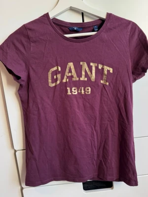 GANT t-shirt. Topp. S.  - Snygg t-shirt från GANT med guldfärgat tryck framtill. T-shirten har rund halsringning och korta ärmar. Storlek S. Perfekt för en avslappnad och trendig look. Sparsamt använd. Hittar inget att anmärka på. Jättefint skick. 
