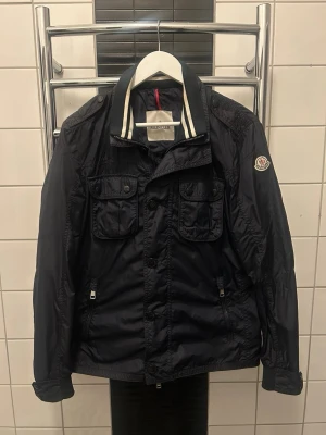Marinblå vindjacka från Moncler - Snygg marinblå vindjacka från Moncler med två bröstfickor med lock, dragkedjeförsedda sidofickor och klassisk krage med vit och svart rand. Blixtlåset har gott sönder men är fixat och fungerar perfekt. Jackan har Moncler-logga på ärmen och är tillverkad i ett lätt, glansigt material som står emot vind. Jackan tillverkas inte mer!                                                                        DM vid intresse🙌🏽