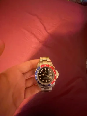 Stilren klocka med röd-blå ring - Säljer en snygg klocka med silverfärgad länk i metall och svart urtavla. Ringen runt urtavlan är tvåfärgad i rött och blått, vilket ger en sportig och cool look. Perfekt accessoar för dig som vill sticka ut lite extra.