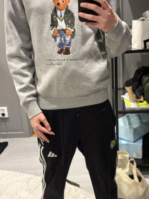 Grå sweatshirt Polo Bear Ralph Lauren - Grå sweatshirt från Ralph Lauren med den ikoniska Polo Bear-trycket framtill. Tröjan har rund halsringning, ribbade muddar vid ärmslut och nederkant samt en avslappnad passform. Perfekt för en casual och trendig look.
