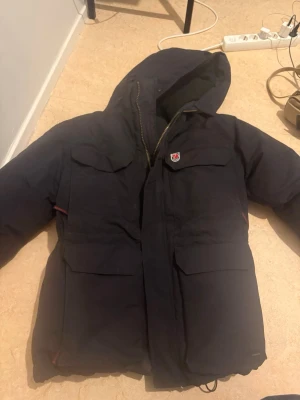 Svart dunjacka från Fjällräven - Säljer en svart dunjacka från Fjällräven med huva och fyra stora fickor framtill. Jackan har klassisk Fjällräven-logga på bröstet och quiltad insida. Perfekt för kalla vinterdagar tack vare det tjocka dunfodret.stor för storlek 