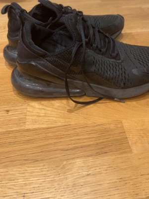 Svarta Nike Air Max 270 - Säljer ett par helsvarta Nike Air Max sneakers med synlig luftbubbla i sulan och meshdetaljer upptill. Skorna har en sportig look med snörning och diskreta loggor på sidan och hälen. Perfekta för dig som gillar streetwear och bekväma skor.