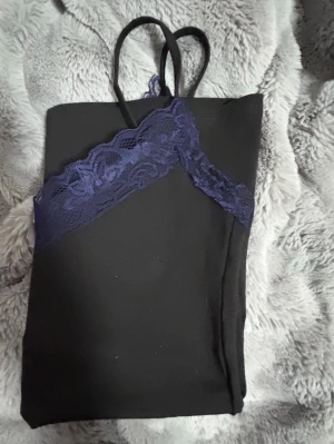 Svart linnetopp med spetskant - Snygg svart linnetopp med tunna axelband och mörklila spetsdetalj längs v-ringningen. Perfekt att styla lager på lager eller bära som den är. Materialet känns mjukt och följsamt, och spetsen ger en extra touch.