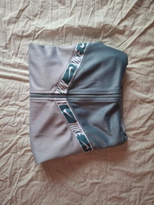 Nike hoodie i grå och blå nyanser - Nike hoodie i tvådelad design med ljusgrå och blågrå färgblock. Dragkedja framtill och huva med snören. Dekorativa band med Nike-logga och swoosh i vitt och grönt över bröstet. Tillverkad i mjukt, stretchigt material som känns sportigt och bekvämt.