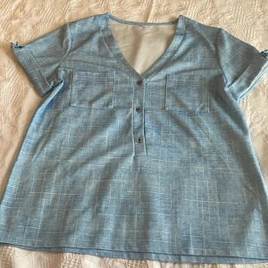 Blå v-ringad blus med fickor - Snygg blå blus med v-ringning och tre knappar framtill. Toppen har korta ärmar med små knyt i ärmsluten och två bröstfickor. Tunt rutigt mönster i vitt ger en fräsch look. Perfekt till jeans eller kjol. Somrig.Någon  mini fläck ärm finns.