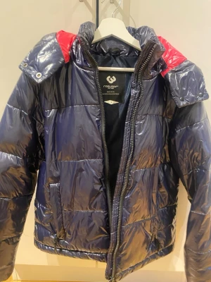 Blå pufferjacka med rosa detaljer  - Snygg blå pufferjacka från Roadwork med glansig finish och kontrasterande röd insida på huvan. Jackan har dragkedja framtill, två sidofickor och en vadderad look som håller dig varm under kalla dagar. Perfekt för dig som vill sticka ut i vinterkylan.