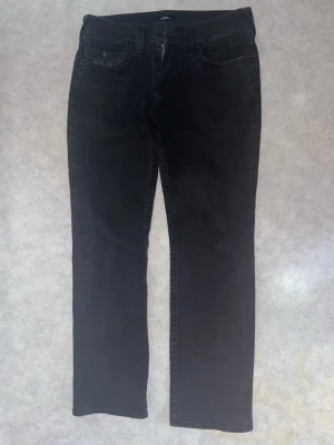 Svarta True Religion jeans - Säljer ett par svarta jeans från True Religion med raka ben och klassiska kontrastsömmar på bakfickorna. Jeansen har fem fickor, bälteshällor och en snygg patch bak i midjan. Materialet är denim med en mjuk känsla och cool look. Pris kan diskuteras