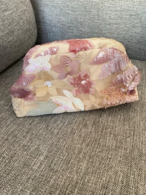 Blommig necessär med paljetter - ALDRIG ANVÄNT!!!  Söt necessär i beige tyg med broderade blommor i rosa, lila och guldiga toner. Utsidan är dekorerad med paljetter som ger ett glittrigt intryck. Väskan har en rektangulär form och svart innerfoder samt guldfärgade detaljer vid öppningen. 
