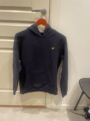 Mörkblå hoodie från Lyle & Scott - Snygg mörkblå hoodie från Lyle & Scott med klassisk gul logga på bröstet. Hoodien har en stor känguruficka framtill och en skön huva. Tillverkad i mjukt bomullsmaterial, perfekt för en chill och avslappnad stil. Storlek 14-15 år skriv om du vill ha mått