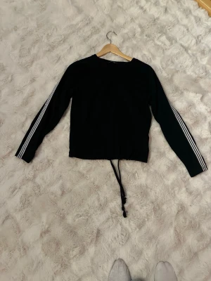 Svart långärmad topp med ränder - Svart långärmad topp med vita ränder längs ärmarna och dragsko i midjan. Sportig och enkel design som passar perfekt till jeans eller träningsbyxor. Mjuk och bekväm