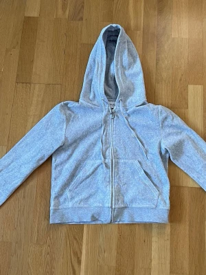 Ljusgrå zip hoodie i velour med huva från Juicy Couture  - Ljusgrå hoodie i velour med dragkedja och stor huva från Juicy Couture i storlek M. Tröjan har långa ärmar, ribbade muddar och en klassisk känguruficka framtill. Materialet är mjukt och bekvämt, perfekt för chill dagar. Enkel och stilren design som passar till det mesta.