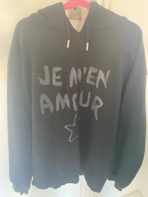 Svart hoodie med text och stjärna - Svart hoodie med huva och dragsko. På framsidan står det 'JE M'EN AMOUR' i glittrig text och en stjärna under. Hoodien har ribbade muddar vid ärmslut och nederkant. Perfekt för en chill och avslappnad stil.