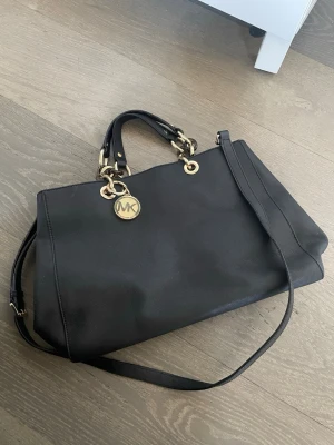 Svart Michael Kors handväska i skinn - Snygg svart handväska från Michael Kors i slätt skinn med guldfärgade detaljer och kedjehandtag. Väskan har både axelrem och dubbla handtag samt en rund MK-logga i metall. Perfekt för dig som vill ha en stilren och rymlig väska med exklusiv känsla. Äkta. Kan finnas små defekter men som knappt syns😊