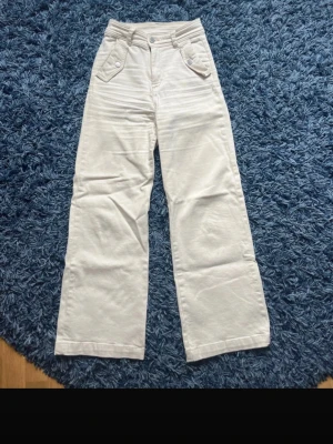 Cream vida jeans med fickdetaljer - Säljer ett par cream jeans med hög midja och vida ben. Jeansen har unika fickor framtill med knappdetaljer och bälteshällor. Materialet är kraftigt och mjukt, perfekt för en avslappnad men trendig look.