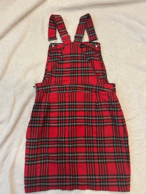 Röd rutigt jumpsuit med hängslen - Cool jumpsuit med klassiskt röd och svart rutigt mönster och justerbara hängslen. Modellen har en rak passform och knappar på axelbanden. Perfekt för dig som gillar en trendig och lekfull stil.