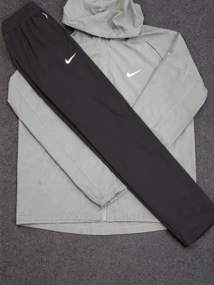 Nike träningsset jacka och byxor - Snyggt träningsset från Nike med en ljusgrå vindjacka med huva och dragkedja samt svarta träningsbyxor med resår i midjan. Båda plaggen har den klassiska vita Nike-loggan. Perfekt för löpning eller gym.