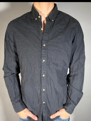 Svart skjorta från Morris - Stilren svart skjorta från Morris med klassisk button-down krage och snygga knappar framtill. Skjortan är långärmad och tillverkad i bomull, perfekt för en clean och enkel look.