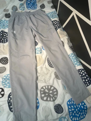 Gråa Under Armour byxor Strolek M - Snygga grå mjukisbyxor från Under Armour med elastisk midja och raka ben. Byxorna har en diskret logga på vänster lår och är tillverkade i ett mjukt och lätt material som passar perfekt för chill eller träning.