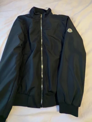 Svart bomberjacka från Moncler - Snygg svart bomberjacka från Moncler med klassisk siluett och ribbade muddar. Jackan har en rak dragkedja framtill och Monclers logotyp på ärmen. Materialet är slätt och ger en clean look som passar till det mesta.