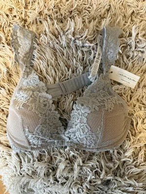 Grå spets-bh från Intimissimi - Snygg grå/vit bh från Intimissimi med breda axelband och vacker spetsdetalj över hela kuporna. Bh:n har ett mjukt mesh-tyg under spetsen och stängs med hyskor bak. Perfekt för dig som gillar romantisk och elegant stil.  Helt ny med lapp