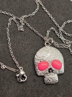 Halsband med dödskalle och neonögon i rostfritt stål  - Unikt halsband med en silverfärgad dödskalleberlock i rostfritt stål dekorerad med blommönster och två neonrosa ögon som lyser i mörkret. Kedjan är tunn och i rostfritt stål och berlocken har coola detaljer som gör den till ett statement-piece. Perfekt för dig som gillar edgy accessoarer.