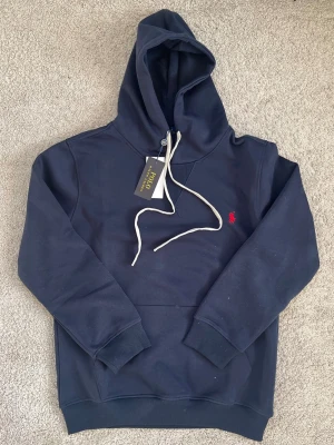 Mörkblå hoodie från Polo Ralph Lauren - Mörkblå hoodie från Polo Ralph Lauren med vit dragsko och klassisk röd logga broderad på bröstet. Hoodien har känguruficka framtill och ribbade muddar vid ärmar och nederkant. Tillverkad i mjuk bomullsblandning för skön komfort. Storleken är M men sitter mer som S tycker jag. Skriv vid intresse 🤗