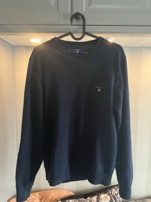 Mörkblå stickad tröja från Gant - Klassisk mörkblå stickad tröja från Gant med rund halsringning och diskret logga broderad på bröstet. Tröjan har ribbade muddar vid ärmslut och nederkant, vilket ger en snygg och bekväm passform. Perfekt till jeans eller chinos.