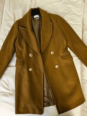 Kamelbrun Ullkappa från Reserved (72% ull) - Snygg ullkappa från Reserved i mycket gott skick. Klassisk dubbelknäppt modell med fin passform och tidlös design. Tillverkad av 72% ull vilket gör den varm och perfekt för vinterdagar. Två fickor framtill och bekväm att bära både för vardagsbruk och finare tillfällen. Storlek L enligt etiketten men passar bättre som M. Säljer då den inte längre används. Klar att användas omedelbart.