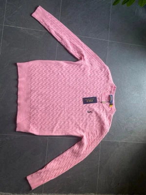 Rosa stickad tröja från Polo Ralph Lauren - Snygg rosa stickad tröja från Polo Ralph Lauren med klassiskt kabelstickat mönster och rund hals. Tröjan har långa ärmar och den ikoniska broderade loggan på bröstet. Perfekt för dig som vill ha en stilren och trendig look.
