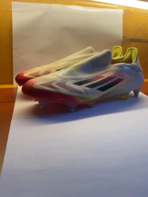 Adidas X Speedflow+ vita fotbollsskor - Säljer ett par Adidas X Speedflow+ fotbollsskor i vitt med röda och gula detaljer samt svarta ränder. Skorna har en strömlinjeformad design utan snörning och är tillverkade i lätt syntetmaterial för maximal fart på planen. Perfekta för dig som vill sticka ut och spela snabbt.