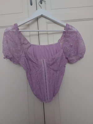 Ljuslila spetskorsett med puffärm - Supersöt ljuslila korsett med puffärmar och detaljer i spets. Modellen har markerade kupor och synliga sömmar framtill för en snygg siluett. Materialet är en blandning av spets och tyg, vilket ger en romantisk och trendig look.