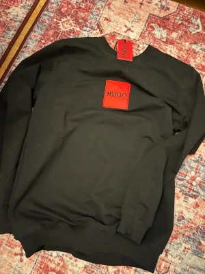 Svart sweatshirt från Hugo - Priset kan diskuteras men jag säljer en svart sweatshirt från Hugo med en stor röd patch och logga på bröstet. Tröjan har rund halsringning, ribbade muddar vid ärmslut och nederkant samt en avslappnad passform. Perfekt för en stilren och modern look. Den är o storlek S och helt sprillans ny med allting kvar.
