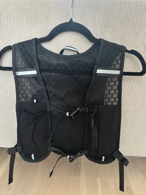 Svart löparväst  - Säljer en svart löparväst med hexagonmönstrad mesh, justerbara remmar och flera praktiska fickor framtill och baktill. Västen har reflekterande detaljer och är tillverkad i lätt, ventilerande material som passar perfekt för löpning eller trail.